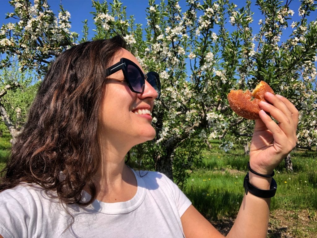 Kate holds a cider donut and smiles at it lovingly. There are apple trees behind her.