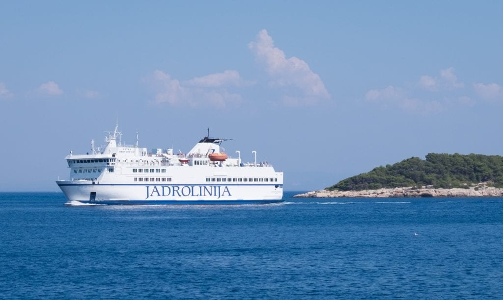 A large white ferry with JADROLINIJA on the side sailing past the edge of an island in Vis.