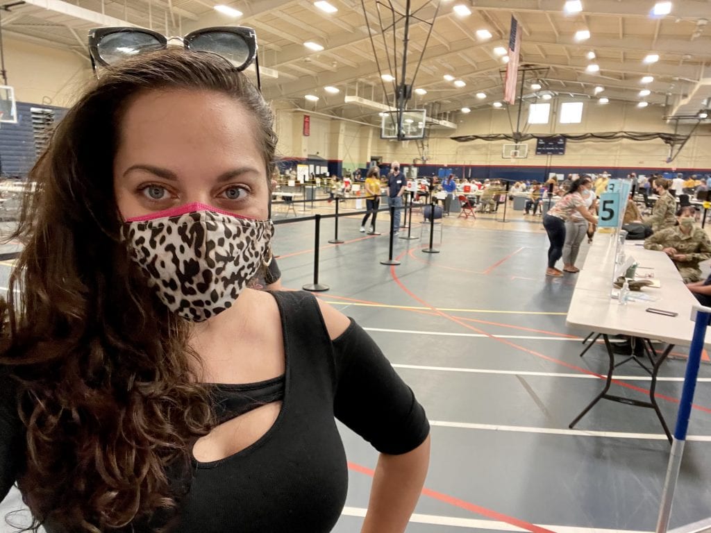 Kate smiling in a leopard print mask on top of a pink mask, wearing a black top with an opening at the shoulder. She stands in a gymnasium where people are getting vaccinated.