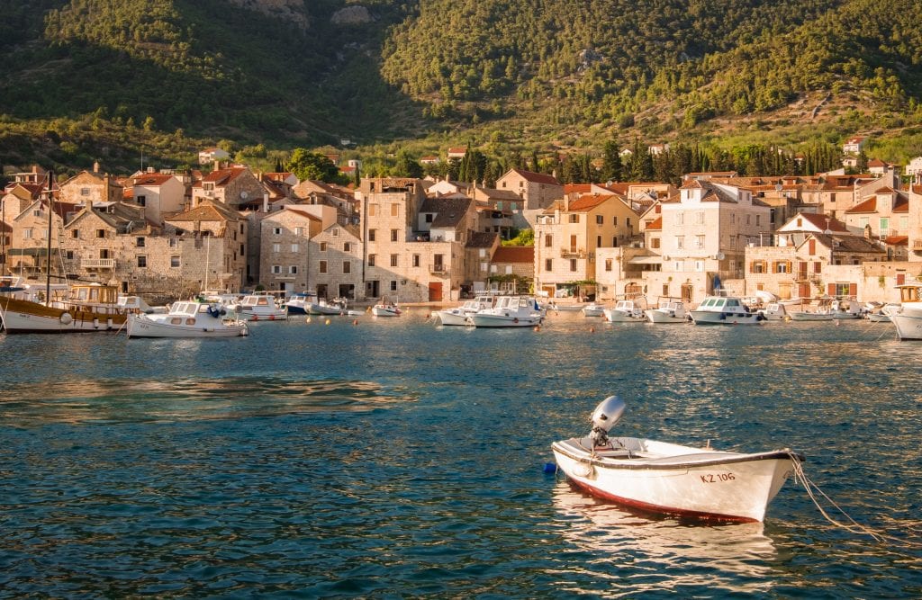 A white stone city on the edge of the bright blue sea, sunset lighting the buildings up in warm shades, a small white rowboat in the foreground.