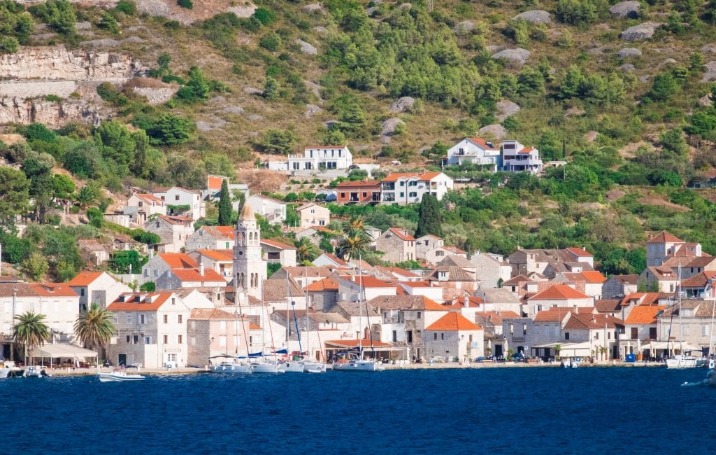 View of Kut from the water: a small town of stone buildings with orange roofs, including a church tower. Some sailboats parked along the water's edge, and the water is dark navy blue.