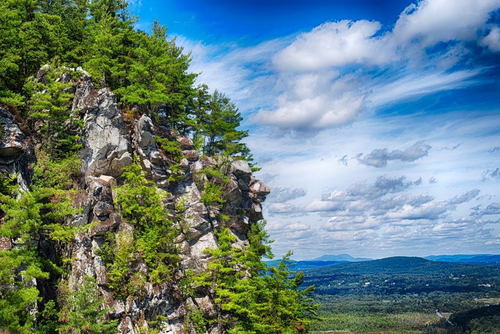 The edge of Monument Mountain, jagged and gray and topped with several evergreen trees, looking out over a cloudy sky and mountains in the distance.