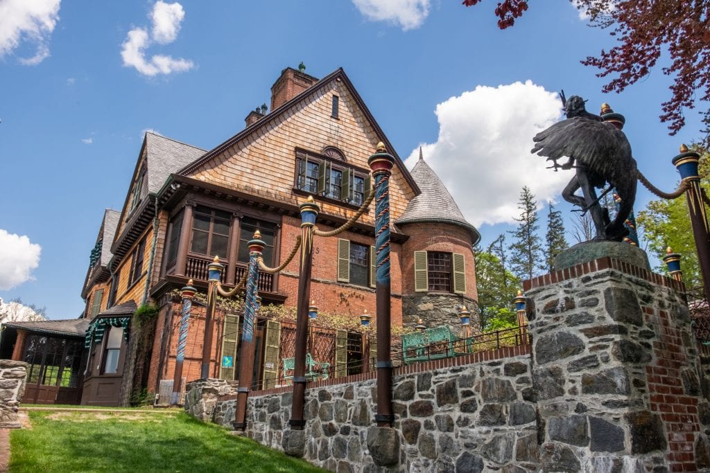 The brown brick-and shingle house of Naumkeag looking over a gray stone patio topped with sculptures and painted wooden poles that look a lot like the wooden poles in the water in Venice.