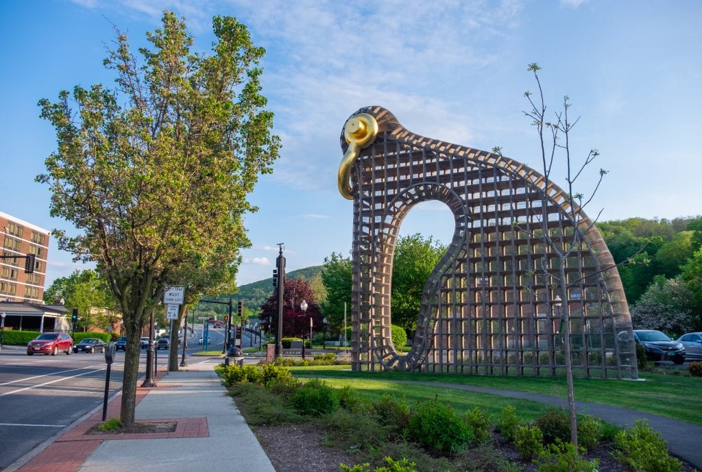 A metal gridded sculpture on the grass that looks a bit like a modern interpretation of a bird, a gold handle serving as the beak and eyeball. It's on a busy street in North Adams.