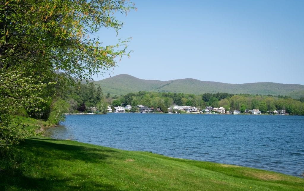 A bright blue lake with white houses on it in the distance, mountains behind them. It is grassy up to the lake's edge.