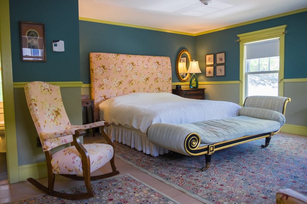 A hotel room filled with 18th century furnishings. Blue walls, a pale peach satin rocking chair, and a long bench at the end of the bed.