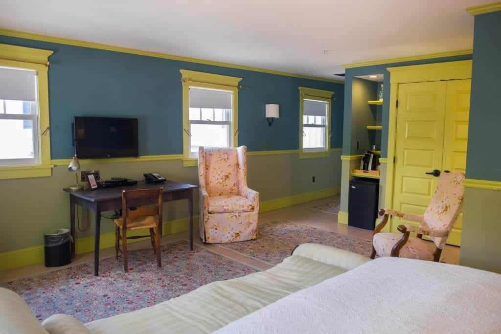 The same room from another angle: you see another chair in the pink and yellow floral print, plus a wooden desk and chair with a much-too-small tv perched above it. A small coffee station and doors to a closet.