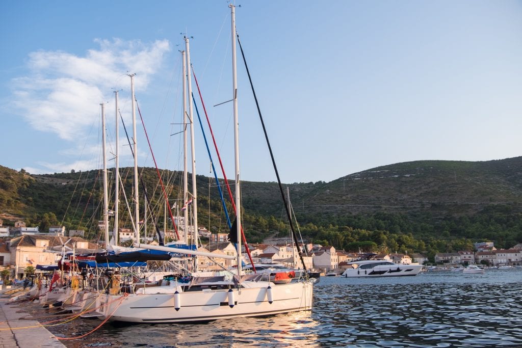 A row of tall white sailboats lined up against the waterfront, each of them with electrical cords plugged into outlets on the cement.