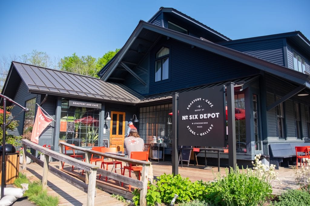 A dark blue restored home with a big sign reading "No Six Depot" in front' also, orange outdoor chairs and people sipping their coffee.