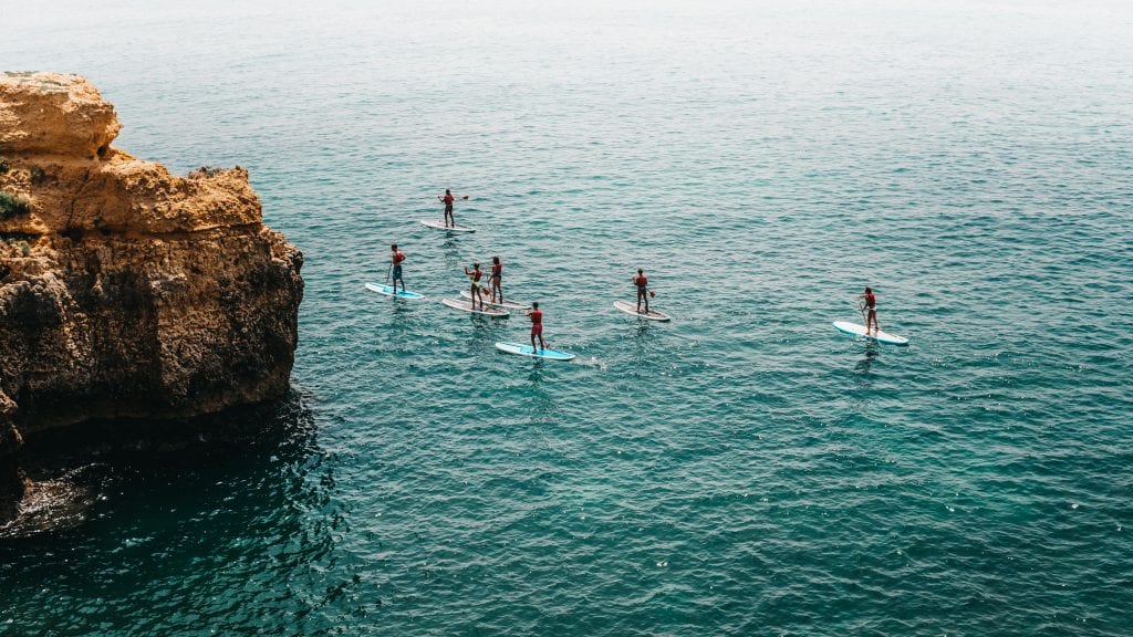 A group of people paddle boarding on the sea, next to a cliff. (This is actually a stock photo from Portugal but nobody knows!)