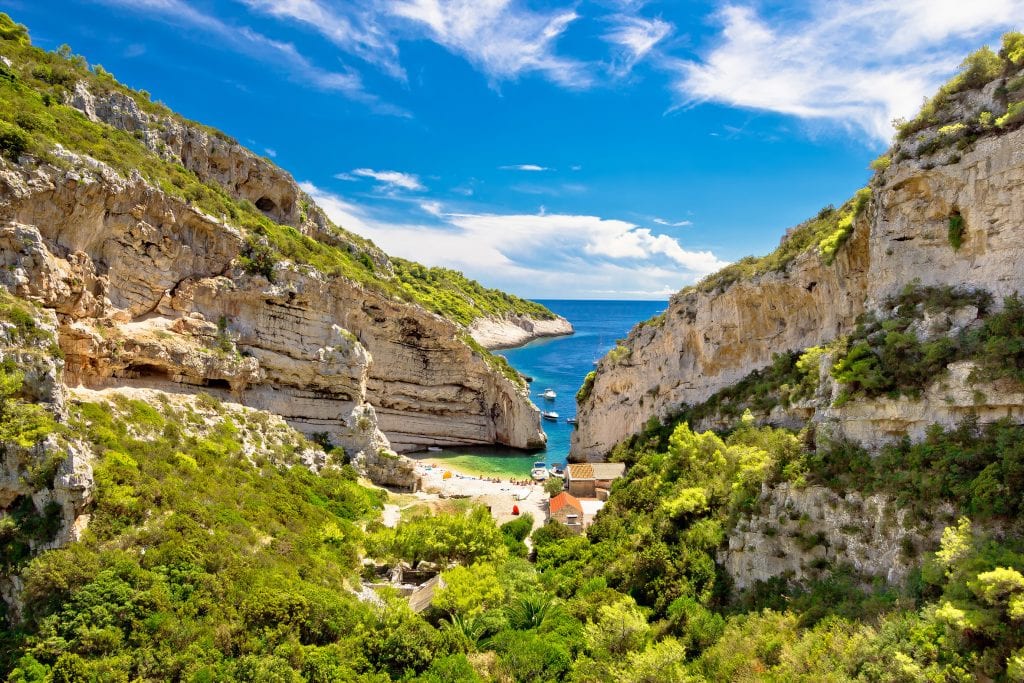 An aerial view heading into Stiniva Bay. You see two rocky cliffs protecting a small white stone beach. It's all surrounded by lush trees.