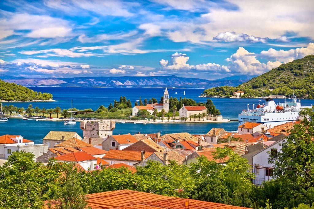 A view over Vis town: you see a small island in the harbor with a church tower, and in the foreground, lots of orange roofs, a ferry, and lots of green trees.