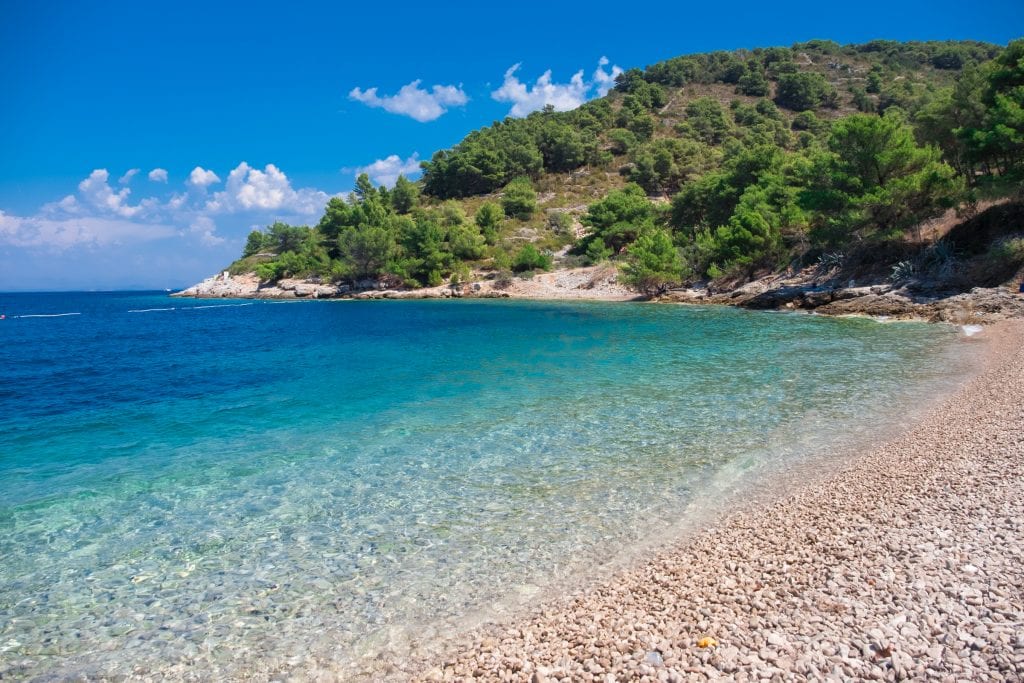 An empty beach (not really, but I photoshopped the people out). Gray pebbles along the beach, and the water is blue-green and nearly clear at the shore. The beach ends and the land rises up, covered with lots of greenery.