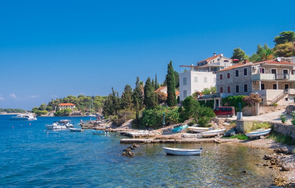 The waterfront in Kut, Vis: several stone buildings along the water's edge, small cement piers jutting into the water, and several small white boats moored in the water.