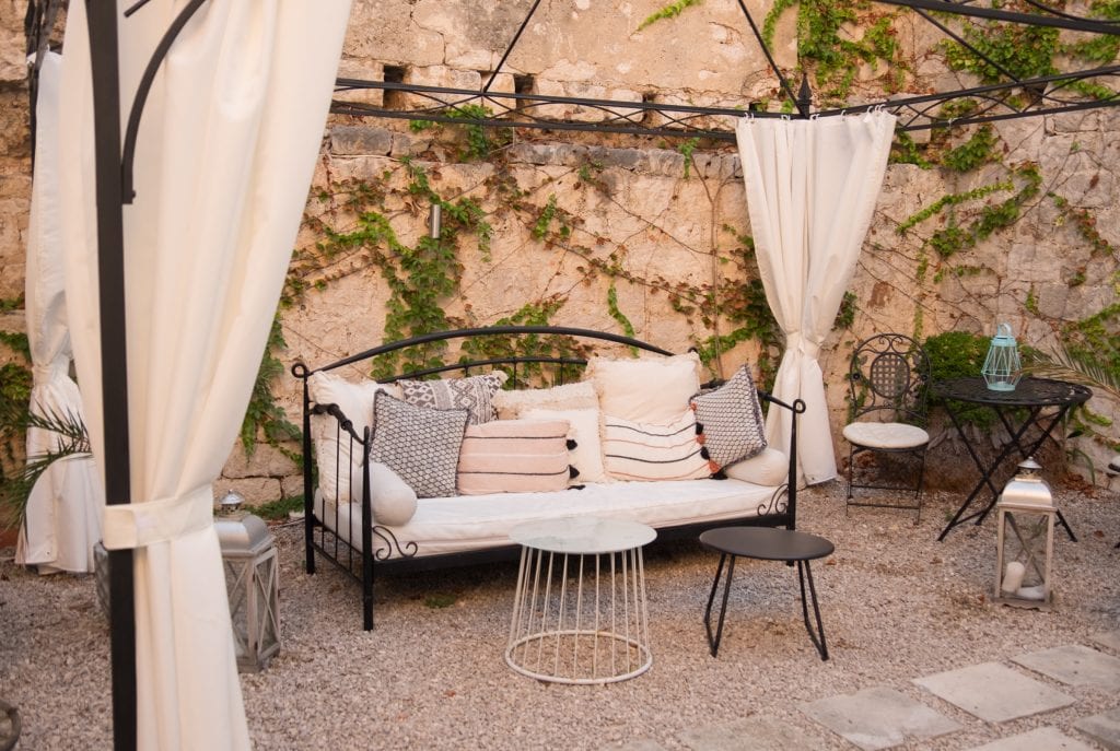 A wrought iron couch covered with beige cushions, surrounded by curtains, in the middle of an outdoor courtyard, the walls covered with ivy.