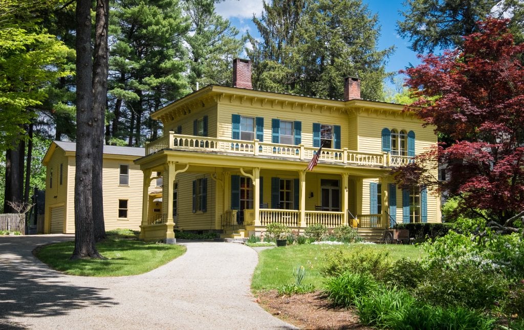 A bright yellow old-fashioned house with a wraparound porch set amongst a wooded area.