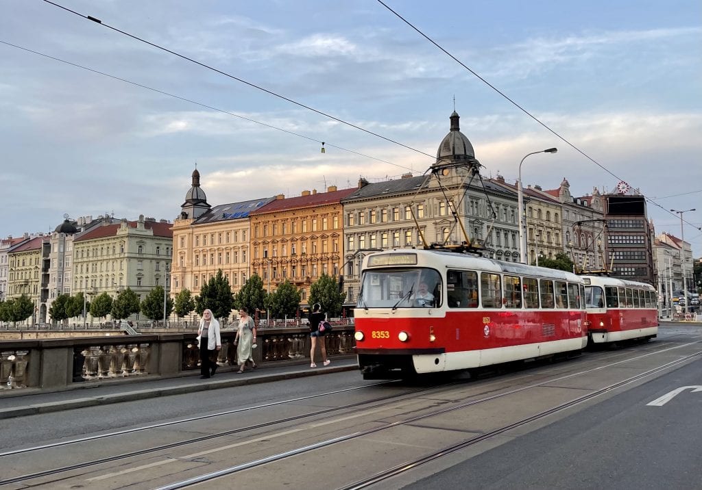 Prague at sunset: you see crenellated buildings on the edge of the river in shades of cream, orange and pale green. In the foreground, a bridge with a red tram going down the street.