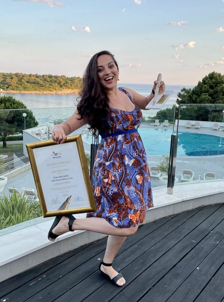 Kate stands in joy holding a gold-framed certificate in one hand and a pen-shaped statue out of stone in the other. She stands in front of the ocean in Croatia. She wears a blue, white and orange dress with shapes of butterflies in the pattern.
