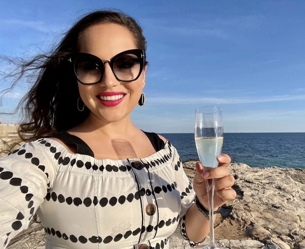 Kate glamour selfie. Kate wears a white off the shoulder Derek Lam top with black polka dots, oversized black Tom Ford sunglasses, gold-edged black Kendra Scott drop earrings, and hot pink lipstick. She holds a glass of champagne and stands on a rocky beach in Croatia.