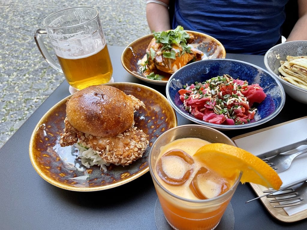 A plate of dishes at a new restaurant, Kro: Charlie has a beer and a roasted chicken drenched in butter chicken sauce; Kate has an orange cocktail and a fried chicken sandwich. Between us is a bowl of kimchi (not REALLY kimchi but kimchi-inspired), bright pink Korean-style marinated cabbage.