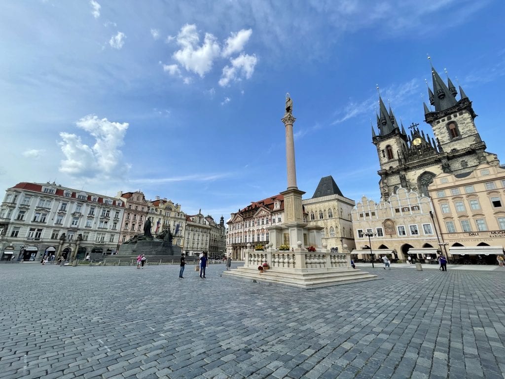 Prague's Old Town Square underneath a blue sky, filled with pastel crenellated buildings and a tall column in the center. Suspiciously empty, as tourists have not yet come back.