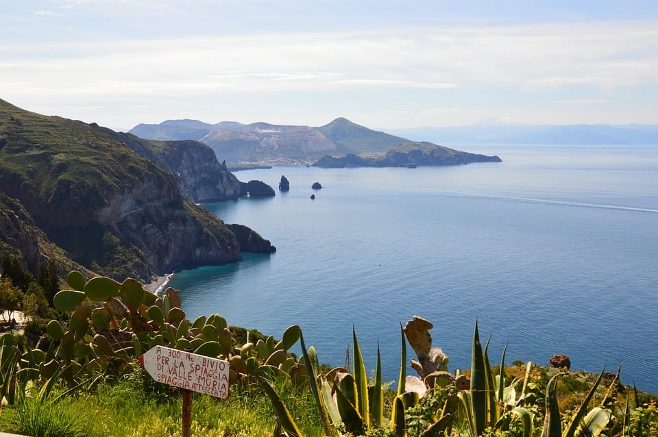 A view overlooking Lipari, one of the Aeolian Islands: A tall, volcanic island coming out of the sea, with lots of tall cliffs. In the foreground, lots of cacti and a hand-painted sign directing to the beach (Spiaggia).