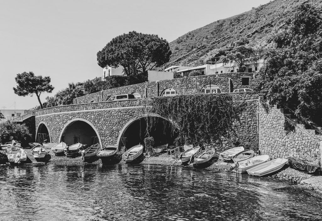 A black and white shot of fishing boats lined up on a tiny beach underneath a bridge, close to the port in Santa Marina Salina.