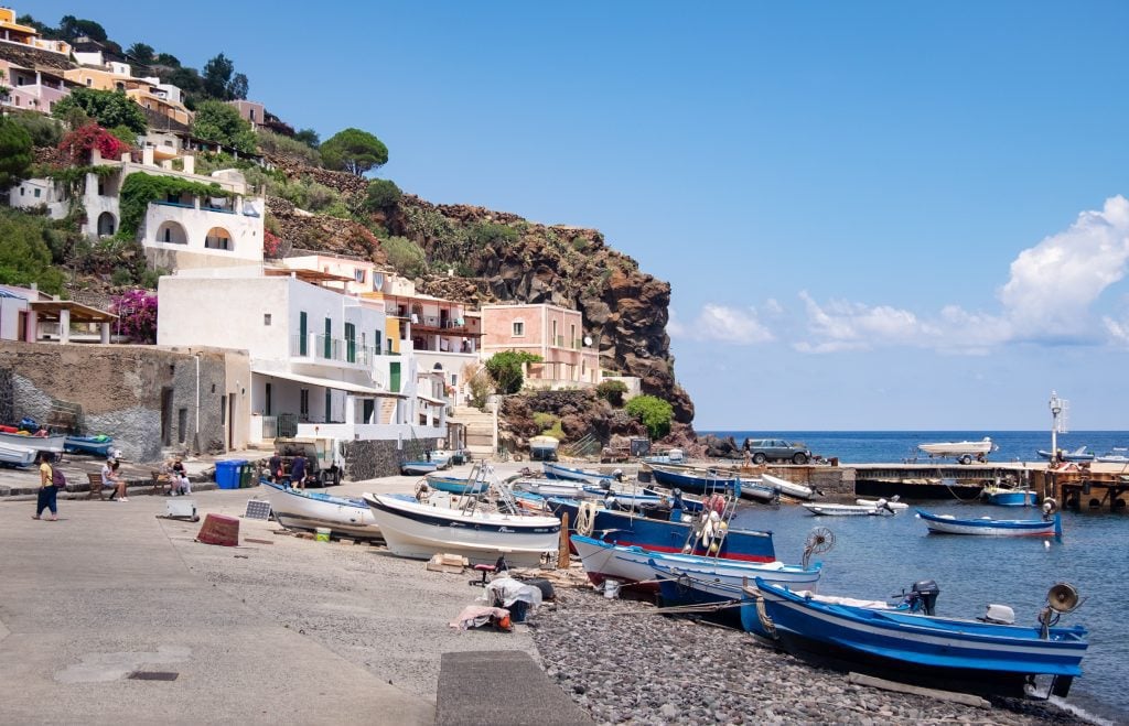 A small fishing village on the island of Alicudi, small wooden boats on the gray stone beach, whitewashed buildings with green doors in the background.