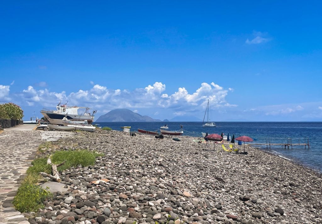 An almost-empty rocky gray beach in Alicudi, one single sailboat in the water.