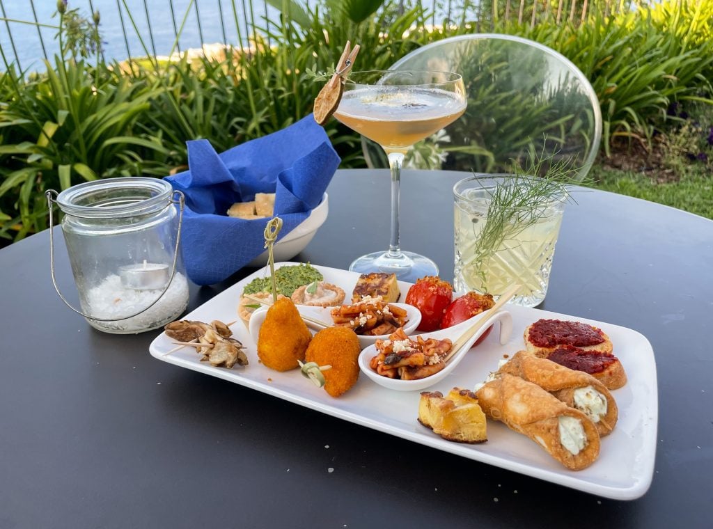 Two cocktails next to an aperitivo plate for two, topped with crostini, spoons filled with pasta alla norma, arancini, stuffed tomatoes, mini cannolis, and other goodies.