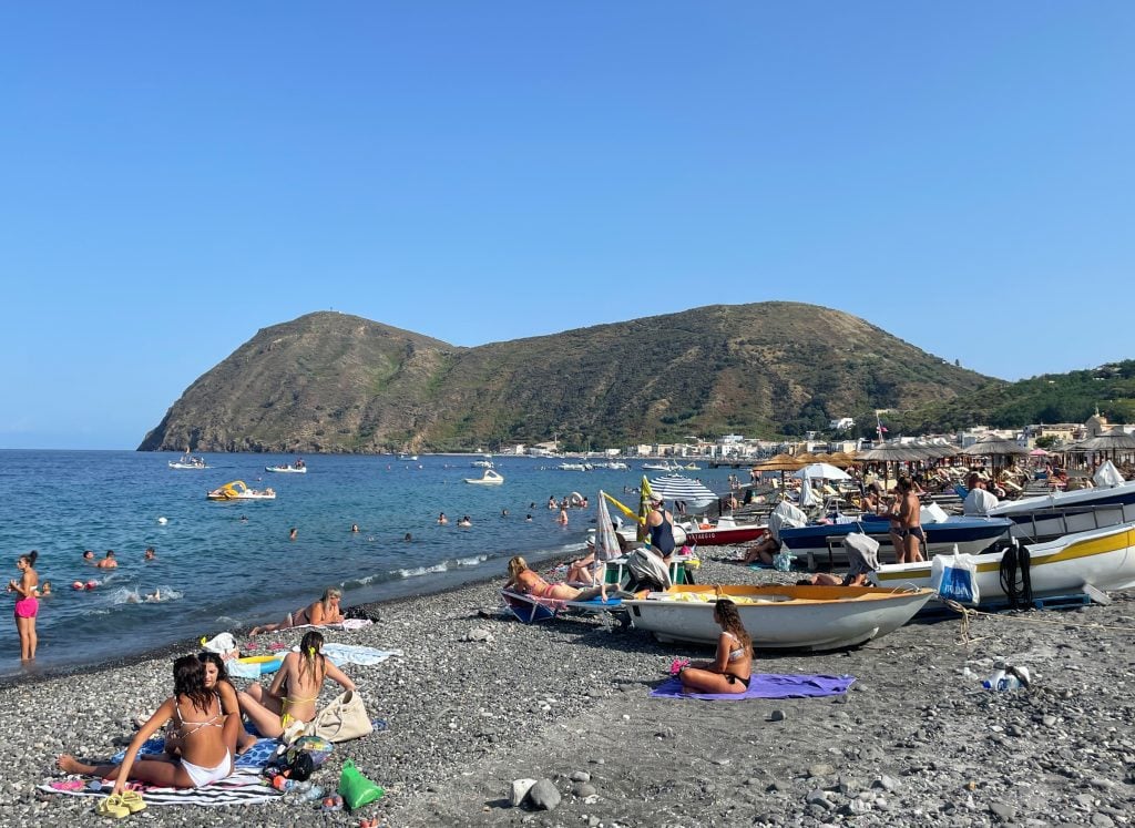 Canneto Beach, a long and overcrowded gray beach with lots of people, kids, floats, boats, and umbrellas.