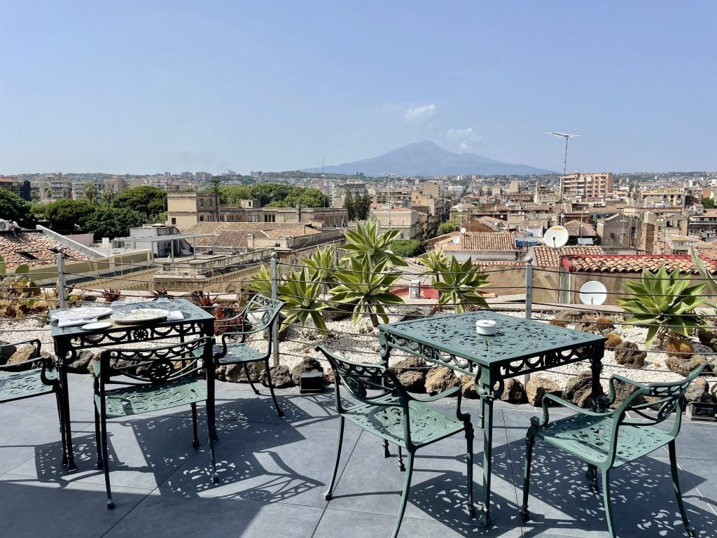 An outdoor terrace with wrought iron tables and chairs overlooking the orange roofs of Catania, with Mount Etna in the distance.