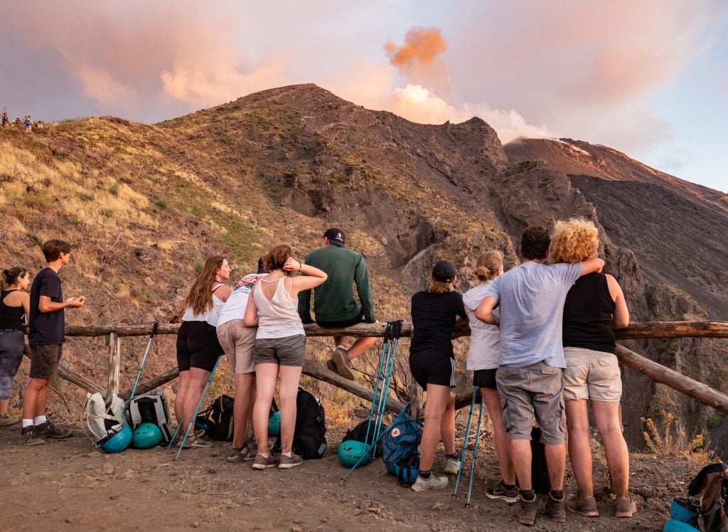 A group of hikers standing in front of a wooden fence, the volcano of Stromboli at sunset emitting pink smoke.