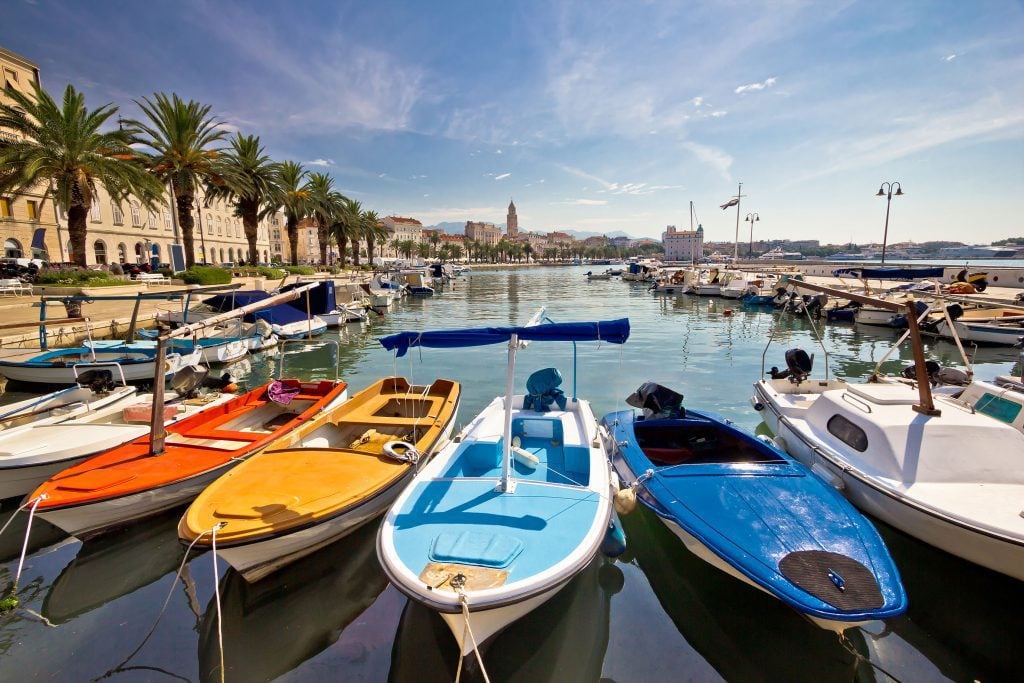 A row of simple painted small boats docked in a harbor with the Split skyline and bell tower in the background.