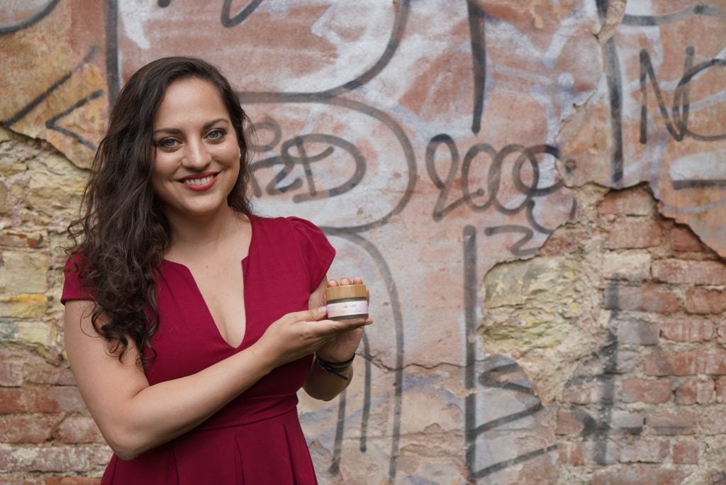 Kate wears a red dress and stands in front of a pink graffitied wall holding a jar of soothing salve.