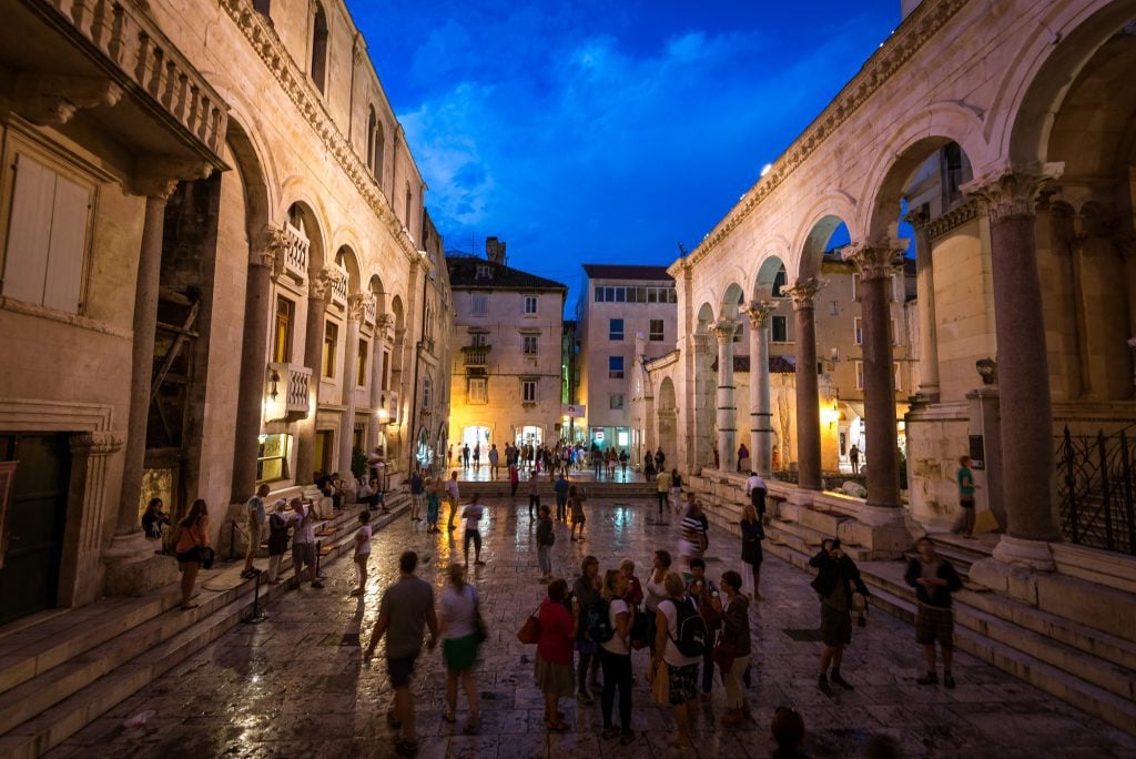 Diocletian's Palace at night: rising columns on stone buildings on each side, with dozens of people walking around in the middle.