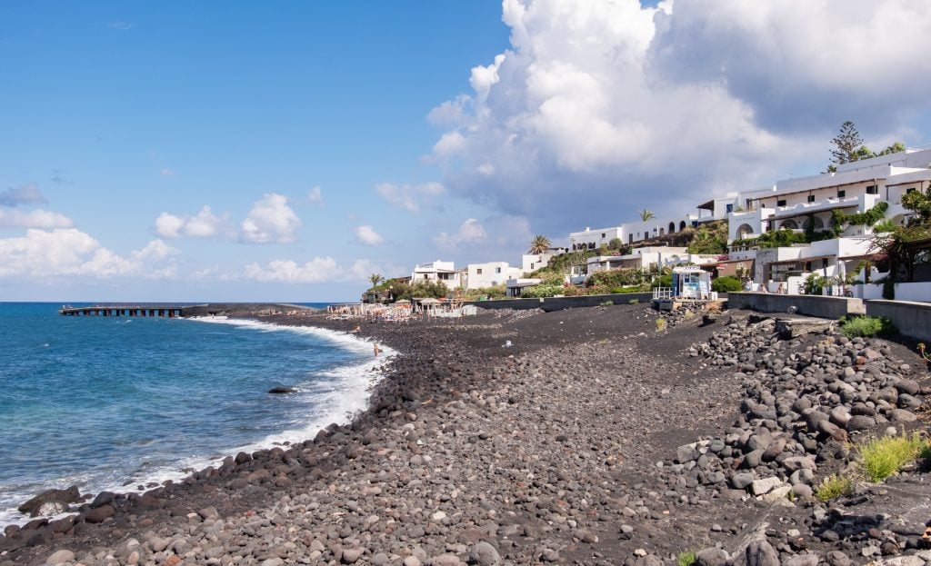 A long black rocky beach in front of the bright blue sea. On shore you see white square-shaped buildings, looking a bit more like Greece than Italy.