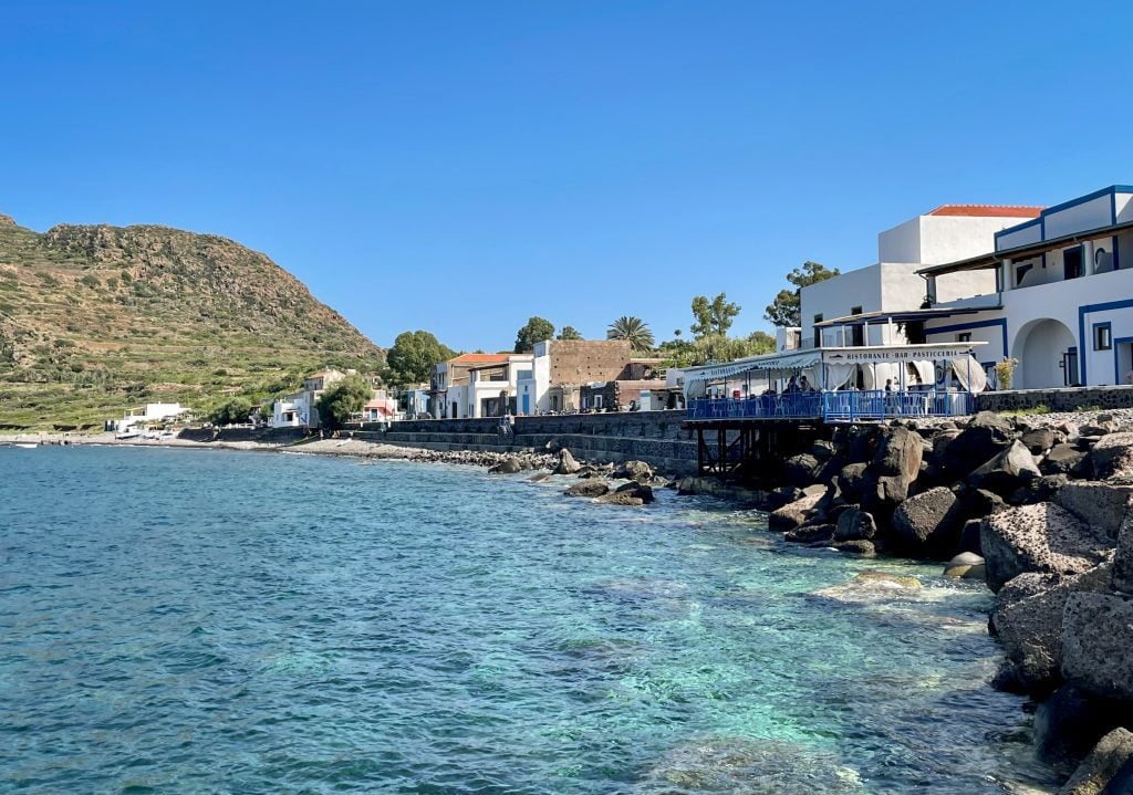 The bright blue coastline of Filicudi, no beaches but a cement wall, and small white buildings on shore.