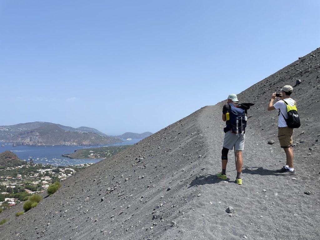 Two men taking a photo while climbing the gray sandy volcano of Vulcano, overlooking islands in the distance.