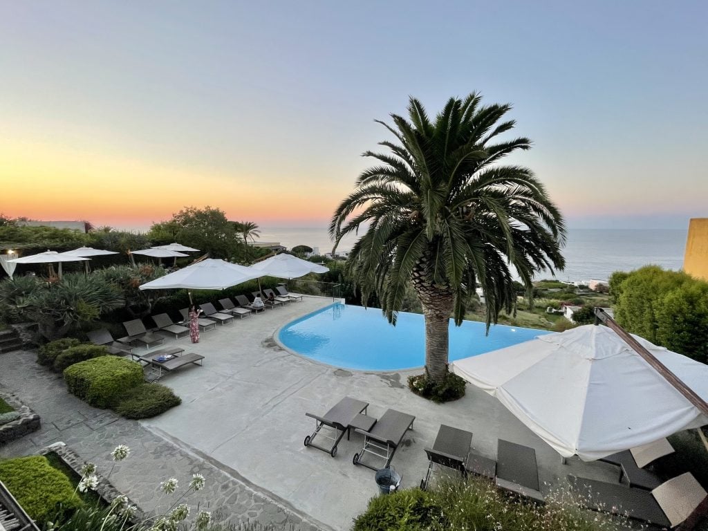 The Hotel Ravesi pool area, with lounge chairs, umbrellas, and palm trees, as seen from above.