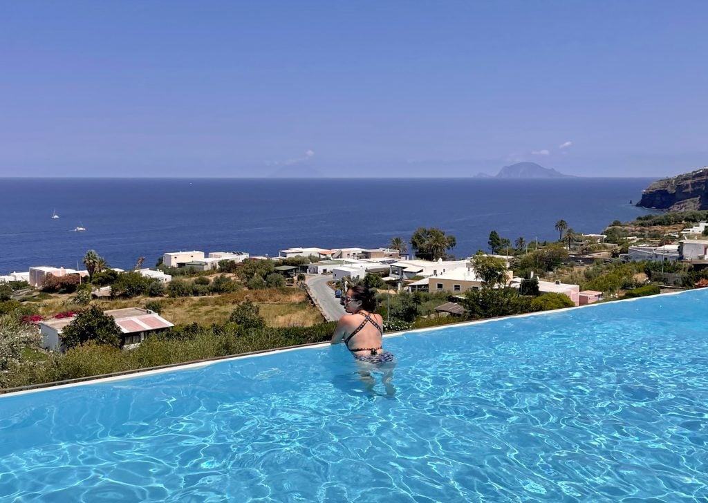 Kate leaning on the edge of the Infinity pool at Hotel Ravesi, overlooking green hills, the ocean, and two islands in the distance.