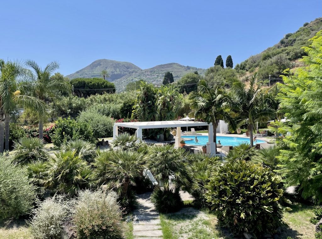 The grounds of Hotel Residence La Giara: a lush green outdoor area with lots of trees and bushes, and in the distance, a small swimming pool filled with kids.