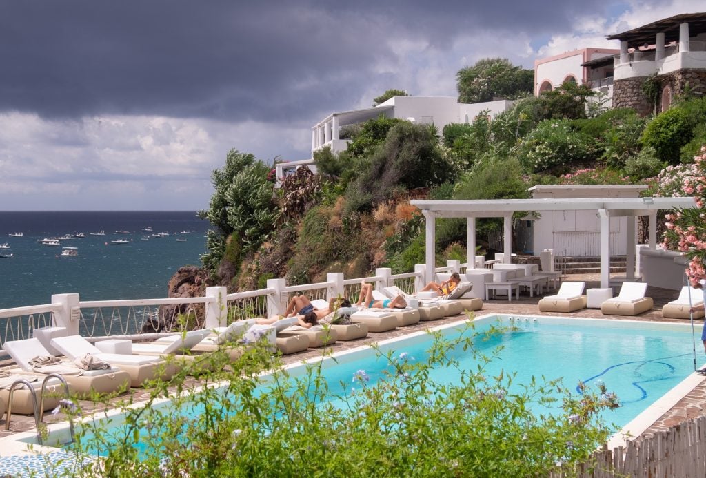 A sneaky photo of a private pool at a hotel on Panarea, guests relaxing on lounge chairs. The sky is gray and looks stormy but blue is appearing along the edges.