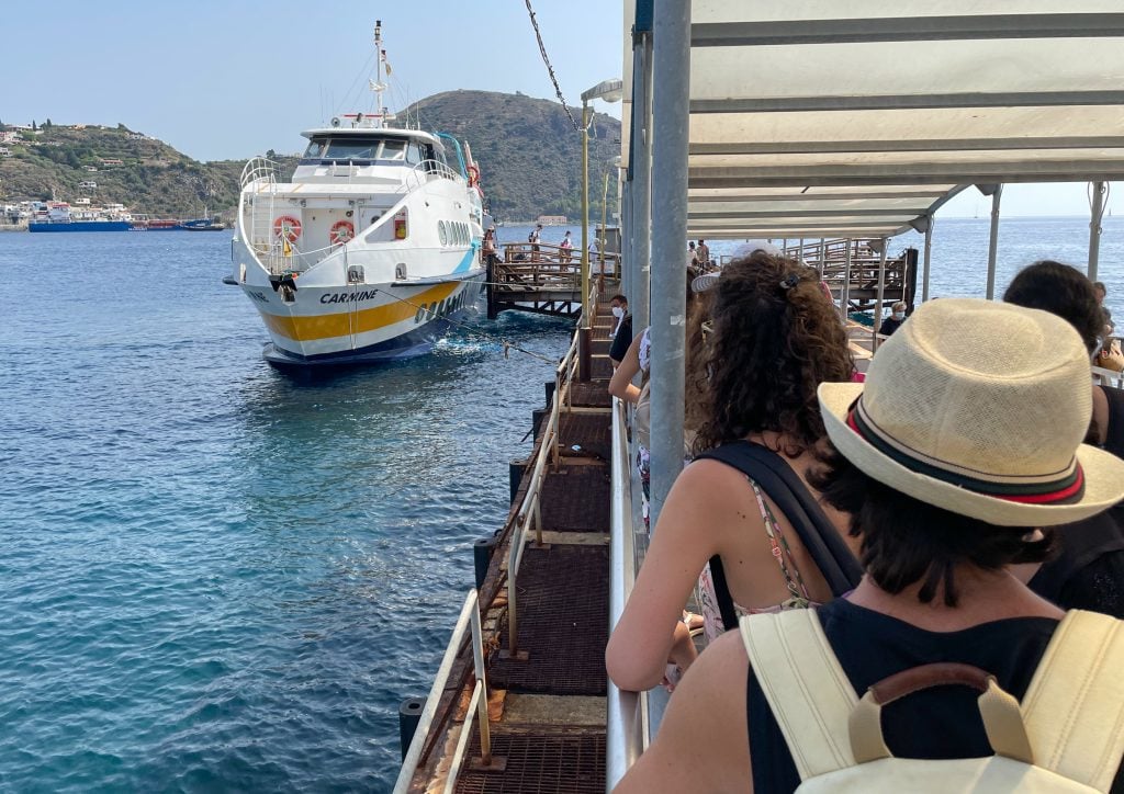 A line of people on a pier waiting to boars a small white hydrofoil named Carmine.