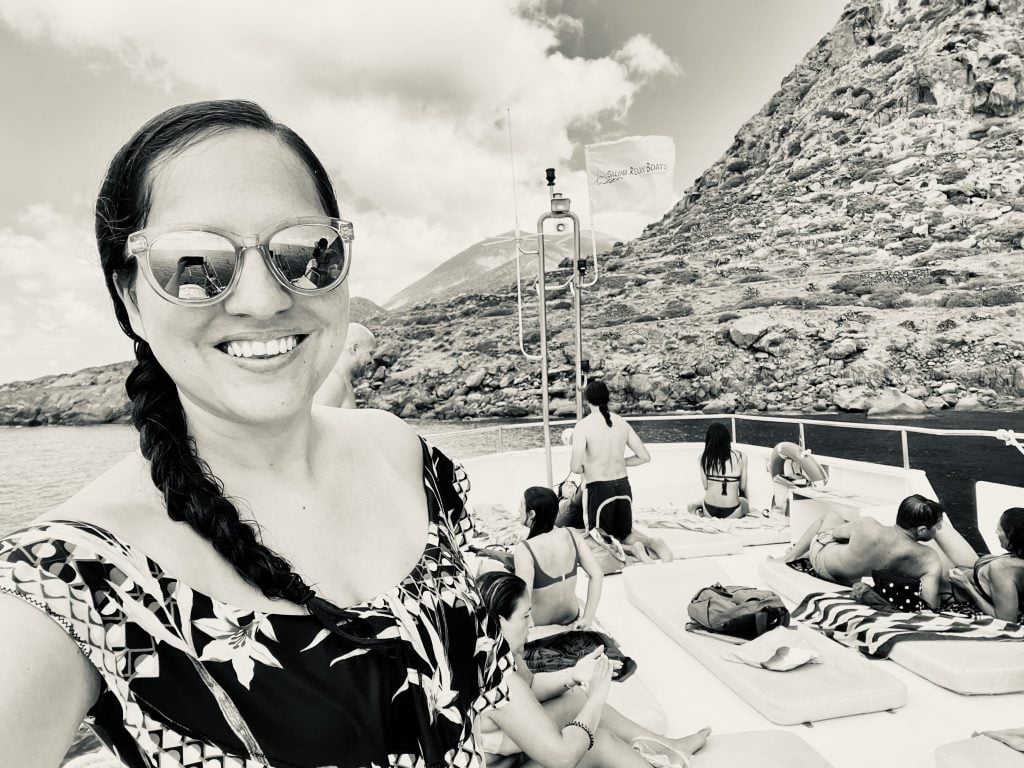 Kate taking a selfie on a boat in Sicily, wearing sunglasses and her hair in a braid. Behind her, people are sunbathing on the boat's top deck; behind that, rocky islands.