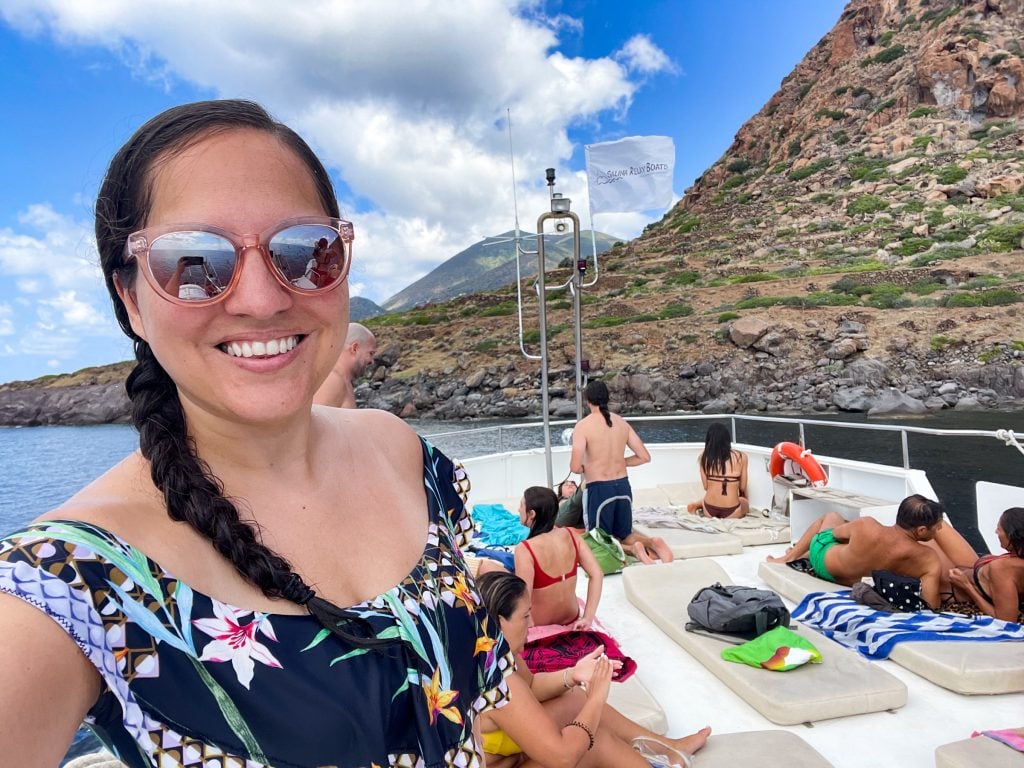 Kate taking a selfie with pink sunglasses, her hair in a single braid, and a tropical-patterned ruffled bathing suit top. She's on the top deck of a boat where people are lounging on mats in their bathing suits. Behind her, you see several islands.