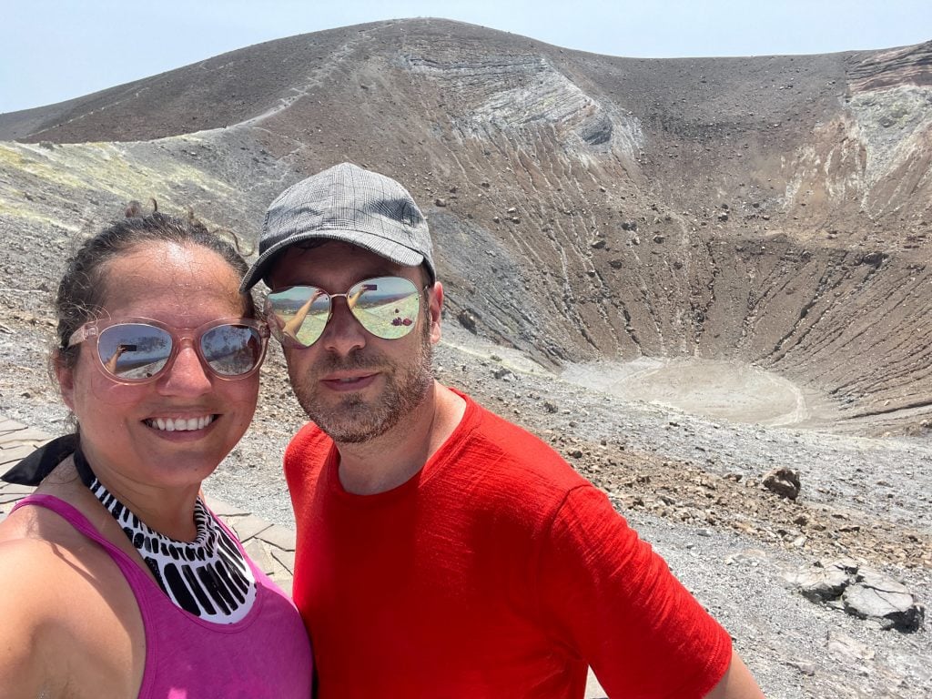 Kate and Charlie taking a sweaty selfie in front of the crater of Vulcano, pale gray-brown, almost like a lunar landscape.