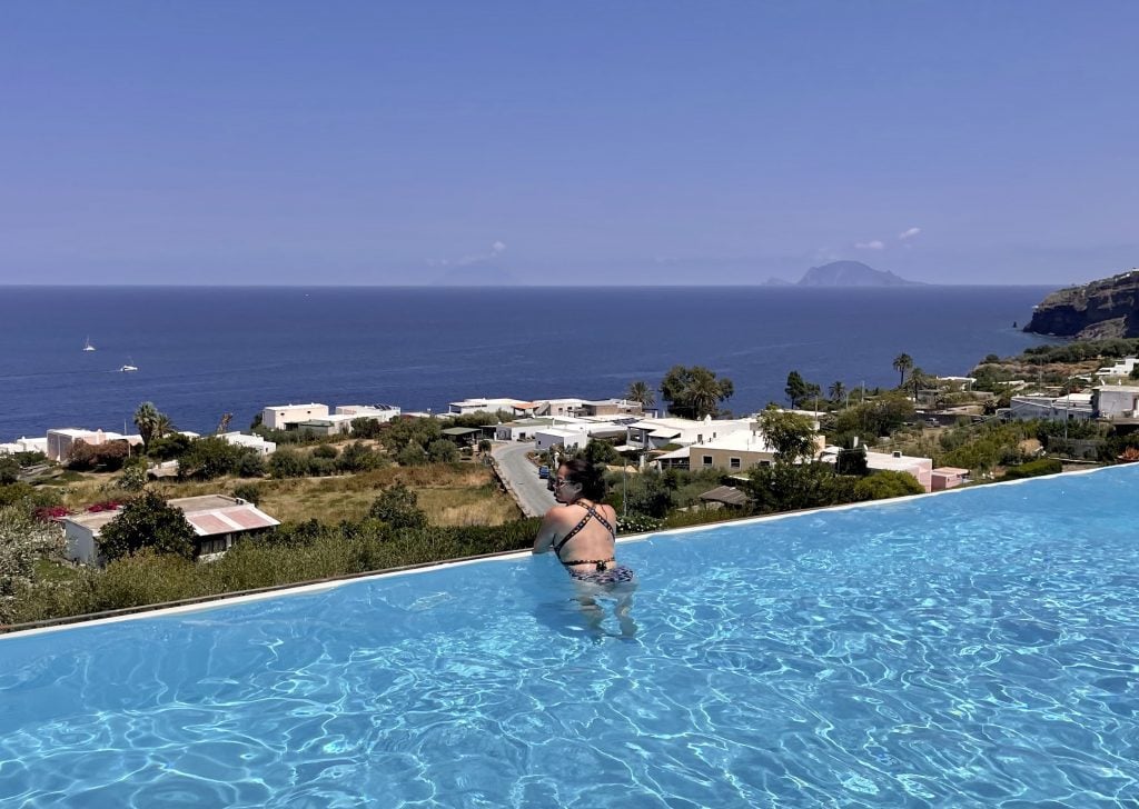 Kate on the edge of the infinity pool at Hotel Ravesi, overlooking the green island landscape with palm trees and white buildings. In the distance in the ocean you can see two islands: Stromboli and Panarea.