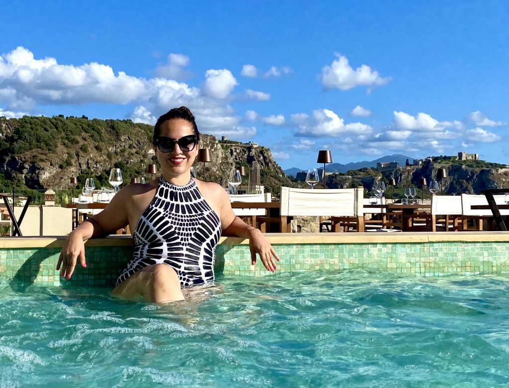 Kate wearing a black and white patterned halter one piece bathing suit and sitting on a pool. Behind her you see rising cliffs and an Italian castle in the far distance.