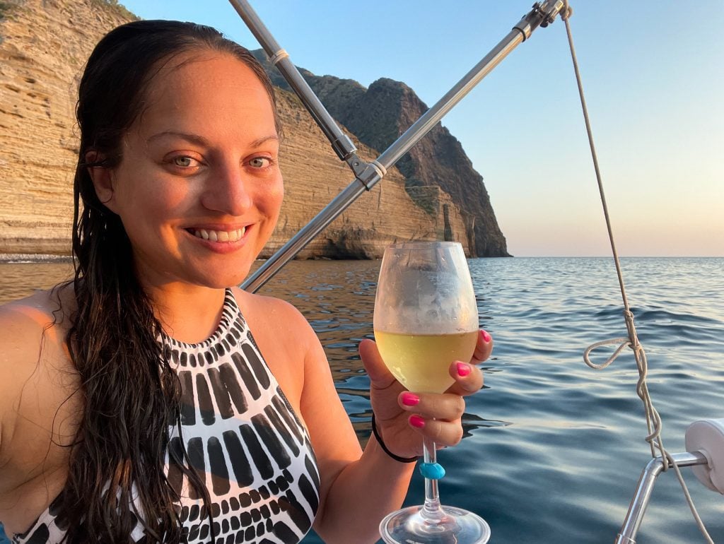 A selfie of Kate smiling with a glass of white wine, wearing a black and white patterned bathing suit. She's on a boat, her hair is long and wet, and you can see Pollara's cliffs behind her.